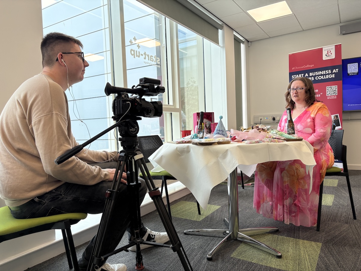 Picture of a videographer conducting an interview with a student. Both are sitting at a table in a classroom setting. 