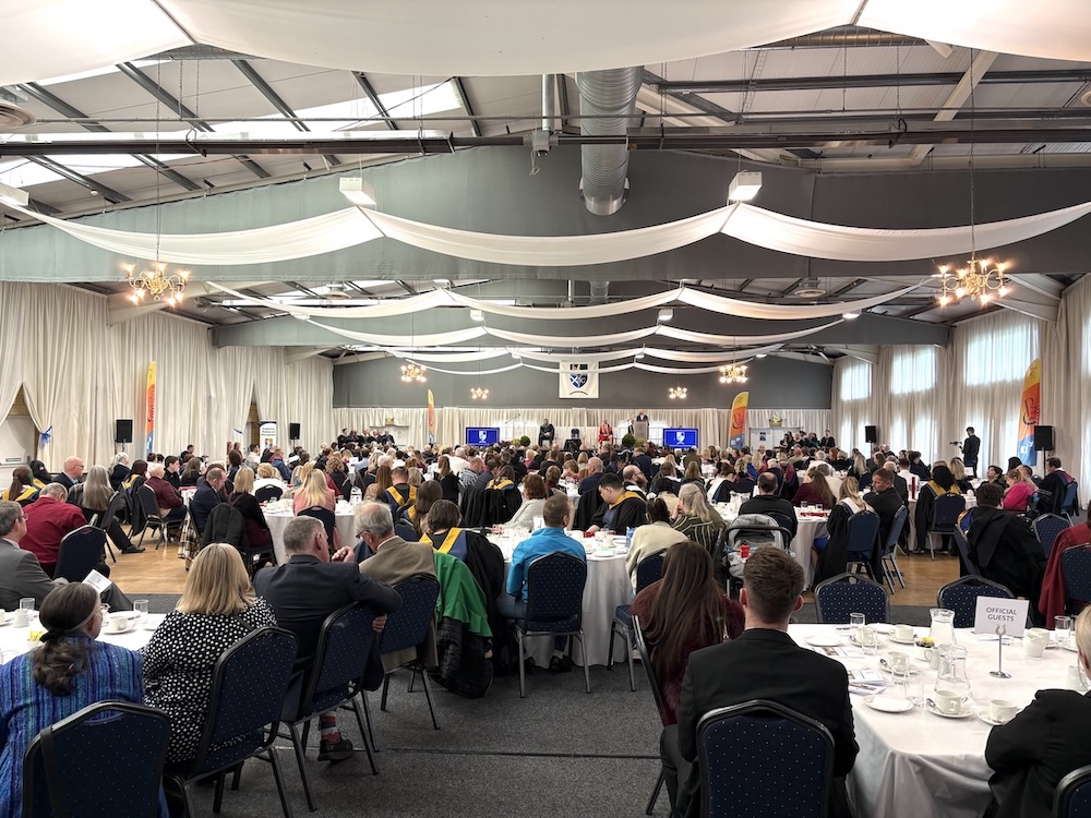 Photograph of the graduation ceremony hall with guests and graduates sitting at round tables. Stage pictured in the background.